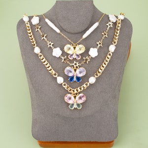 May include: A layered necklace with a gold chain, pearl accents, and three butterfly charms. The butterflies are made of colorful rhinestones and have a layered, dimensional look.