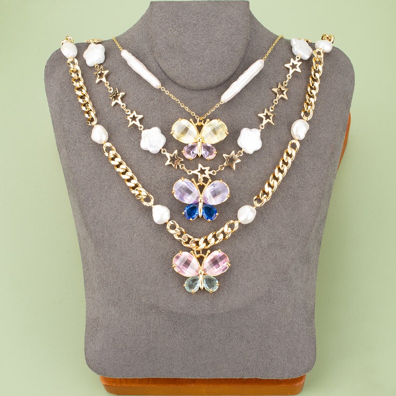 May include: A layered necklace with a gold chain, pearl accents, and three butterfly charms. The butterflies are made of colorful rhinestones and have a layered, dimensional look.