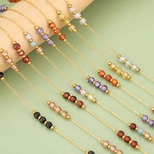 May include: A selection of gold-coloured chain necklaces, each featuring a string of colourful, faceted beads. The beads are in shades of red, purple, yellow, and clear. The necklaces are displayed against a light green background.