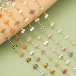 May include: Close-up of several strands of colorful beaded chain. The beads are in various colors, including white, pink, orange, and purple. The beads are strung on gold-colored chains. The background is a light green color.