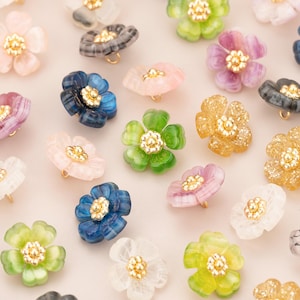 Puede incluir: Surtido de botones de resina en forma de flor de varios colores con detalles dorados. Los botones están dispuestos sobre una superficie blanca.