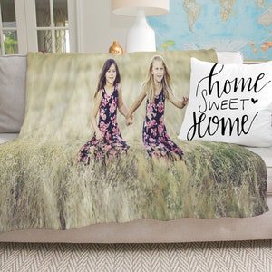May include: A throw blanket and pillow on a beige sofa. The blanket features a photo of two girls in floral dresses in a field of tall grass. A white pillow displays the words "home sweet home" in black script.