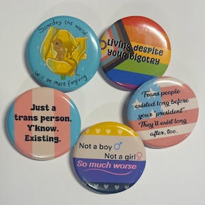 Peut inclure: Cinq badges ronds avec différents messages et designs. Les badges sont colorés et présentent une variété de thèmes, notamment l'identité de genre, la justice sociale et l'activisme politique. Le texte sur les badges comprend des phrases comme "Vivre malgré votre bigoterie", "Juste une personne trans. Tu sais. Exister", et "Les personnes trans existaient bien avant votre "président". Elles existeront longtemps après, aussi."