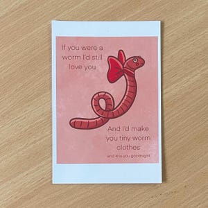 May include: A greeting card with a pink background and a cartoon worm illustration. The worm has a red bow and is wearing tiny clothes. Text on the card reads, "If you were a worm I'd still love you. And I'd make you tiny worm clothes and kiss you goodnight."