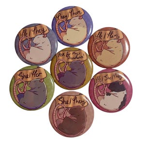 May include: A collection of seven round buttons with illustrations of sleeping rats. Each button has a different color background and text indicating pronouns such as "He/They", "She/Her", and "They/Them". The buttons are a mix of colors.