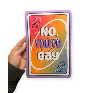 Pode incluir: Um cartão retangular com uma borda roxa e um fundo gradiente de arco-íris. O cartão apresenta o texto "NO YOU'RE GAY" em branco e roxo, com setas invertidas e detalhes de estrelas. O cartão é segurado por uma mão.