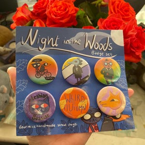 May include: A set of six round pinback buttons featuring characters from the video game "Night in the Woods". The buttons are arranged in a grid on a blue background with the text "Night in the Woods Badge Set" and "fourmice handmade. straw. page".