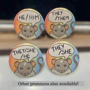 Puede incluir: Cuatro botones redondos con un fondo de arcoíris y una rata de dibujos animados. Cada botón tiene un conjunto diferente de pronombres escritos en un globo de diálogo sobre la rata. Los pronombres son "HE/HIM", "THEY/THEM", "THEY/SHE/HE" y "THEY/SHE". El texto "¡Otros pronombres también disponibles!" está escrito debajo de los botones.