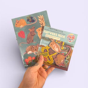 Könnte beinhalten: Eine Hand hält zwei Stickerpackungen. Eine Packung zeigt Illustrationen von Erntemäusen mit Beeren und Weizen. Die andere Packung ist mit "Critter Club Gloss Sticker Collection Harvest Mice" beschriftet. Die Aufkleber sind wasser- und wetterfest.