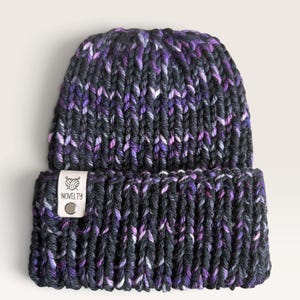 May include: A chunky knit beanie in a variegated blend of dark charcoal gray, deep purple, and bright white yarn. The hat features a wide folded cuff with a rectangular light-colored leather tag embossed with the word NOVELTY and a yarn ball icon.