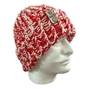 Puede incluir: Un gorro de punto rojo y blanco con un diseño acanalado. El gorro tiene una pequeña etiqueta rectangular que dice "NOVELTY". El gorro se muestra en una cabeza de maniquí blanca.