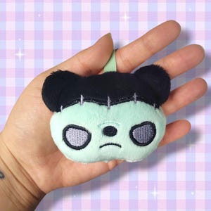 May include: A plush Frankenstein bear keychain with a mint green face, black hair, and stitched details. The bear has embroidered gray eyes and a small black nose. A green loop is attached for hanging.