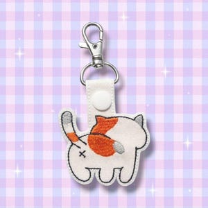 Op de afbeelding: Een sleutelhanger met een geborduurd kattenkont-ontwerp. Het ontwerp toont een witte kat met oranje en grijze markeringen en een zwarte omtrek. De sleutelhanger heeft een witte band met een drukknoopsluiting en een zilveren karabijnhaak.