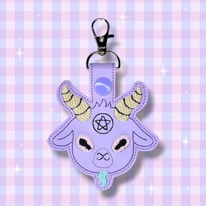 May include: A lavender keychain featuring an embroidered goat head with a pentagram, horns, and an infinity symbol. The keychain has a silver clasp and a matching lavender strap. The background is a checkered pattern in shades of purple and white.