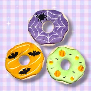 以下が含まれることがあります： ハロウィーンをテーマにした3つのぬいぐるみドーナツ。1つはクモの巣とクモが付いた紫、もう1つはコウモリが付いたオレンジ、最後はカボチャとキャンディーコーンが付いた緑です。ドーナツには白いフロスティングトリムが付いています。
