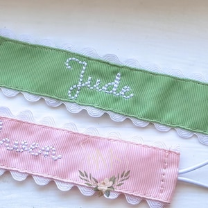 May include: Two pacifier clips with metal clasps. One clip is light green with the name "Jude" embroidered in white. The other is pink with the name "Grace" and floral embroidery. Both have white lace trim and white elastic loops.