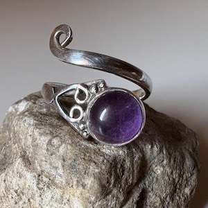 May include: A silver-toned ring with a large, round, purple gemstone. The ring has an open design with a decorative swirl and is displayed on a textured rock. The gemstone is a deep, rich purple color.