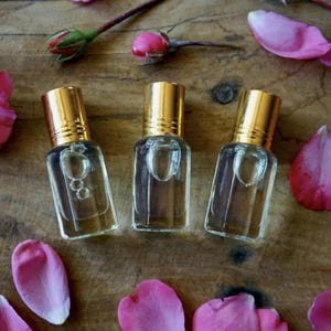 May include: Three small, clear glass perfume bottles with gold caps are arranged on a wooden surface, surrounded by pink rose petals, buds, and a full bloom. The bottles are filled with a clear liquid.