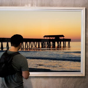 Puede incluir: Fotograf&iacute;a enmarcada de un muelle que se extiende hacia el oc&eacute;ano al atardecer. El cielo transita del amarillo al naranja, con la silueta del muelle y el reflejo del agua visibles. Una persona est&aacute; mirando la fotograf&iacute;a enmarcada.