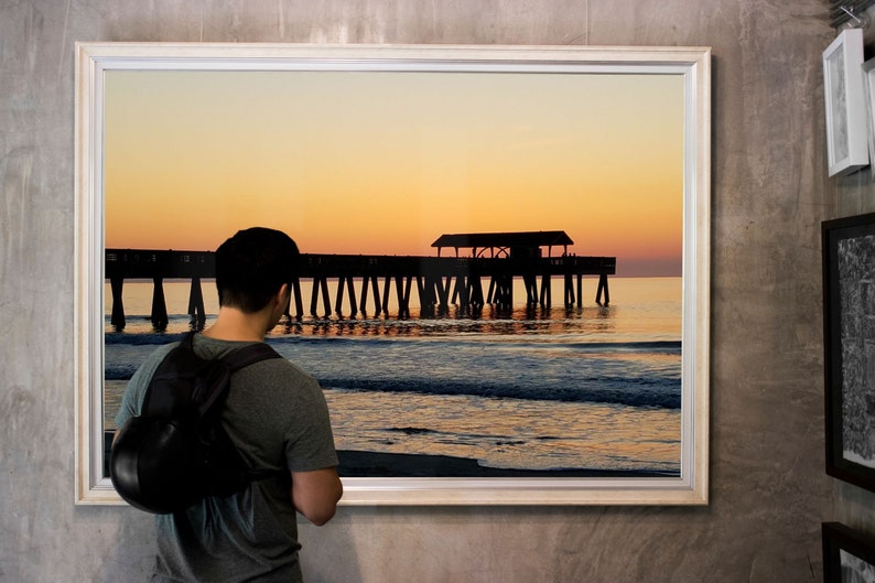 Puede incluir: Fotograf&iacute;a enmarcada de un muelle que se extiende hacia el oc&eacute;ano al atardecer. El cielo transita del amarillo al naranja, con la silueta del muelle y el reflejo del agua visibles. Una persona est&aacute; mirando la fotograf&iacute;a enmarcada.