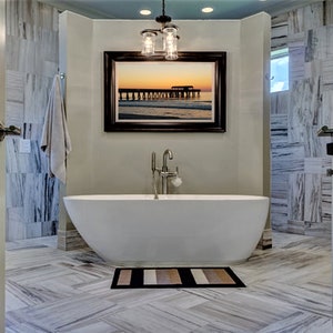 Puede incluir: Un ba&ntilde;o moderno con una ba&ntilde;era ovalada blanca, una foto enmarcada de un muelle al atardecer y una alfombra de ba&ntilde;o a rayas. Las paredes y el suelo presentan un patr&oacute;n similar al m&aacute;rmol. Se accede a la habitaci&oacute;n a trav&eacute;s de dos puertas blancas abiertas.