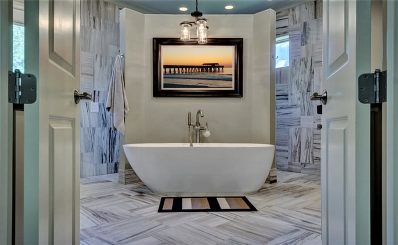 Puede incluir: Un ba&ntilde;o moderno con una ba&ntilde;era ovalada blanca, una foto enmarcada de un muelle al atardecer y una alfombra de ba&ntilde;o a rayas. Las paredes y el suelo presentan un patr&oacute;n similar al m&aacute;rmol. Se accede a la habitaci&oacute;n a trav&eacute;s de dos puertas blancas abiertas.