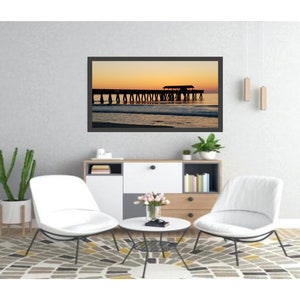 Puede incluir: Una fotograf&iacute;a enmarcada de un muelle al atardecer, con el cielo pasando de naranja a amarillo. La silueta del muelle es visible contra el cielo colorido. La fotograf&iacute;a se exhibe en un sal&oacute;n moderno con sillas blancas y una alfombra estampada.