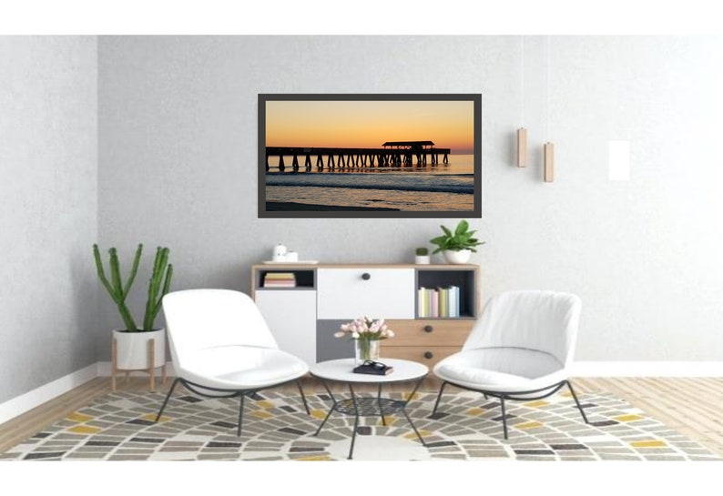 Puede incluir: Una fotograf&iacute;a enmarcada de un muelle al atardecer, con el cielo pasando de naranja a amarillo. La silueta del muelle es visible contra el cielo colorido. La fotograf&iacute;a se exhibe en un sal&oacute;n moderno con sillas blancas y una alfombra estampada.