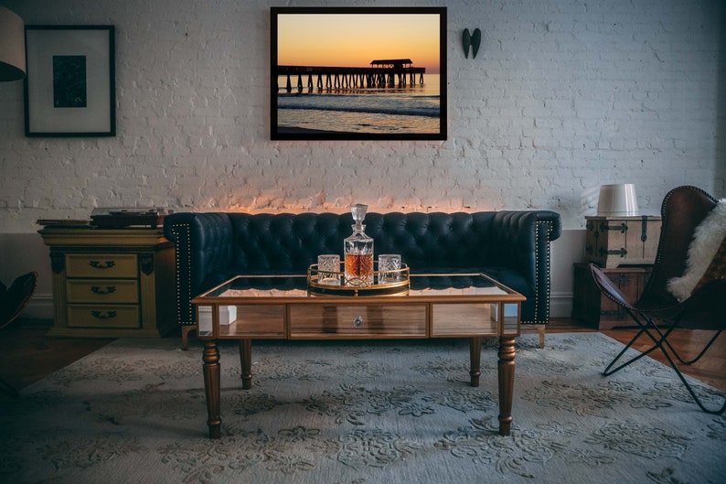 Puede incluir: Un sal&oacute;n con un cuadro enmarcado de un paisaje de puesta de sol. Un sof&aacute; de cuero azul oscuro, una mesa de centro de madera con tapa de espejo y un juego de decantador est&aacute;n en primer plano. La habitaci&oacute;n tiene una alfombra y una pared de ladrillo blanco.