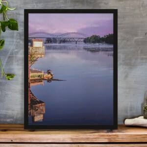 May include: Framed landscape print featuring a serene river scene at dawn. The image showcases a bridge in the distance, a small structure on the water's edge, and the reflection of the scene in the calm water. The sky is a blend of pink and purple hues.