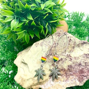 Könnte beinhalten: Ein Paar silberne Ohrhänger mit einem Regenbogenherz-Anhänger und einem silbernen Marihuana-Blatt-Anhänger.