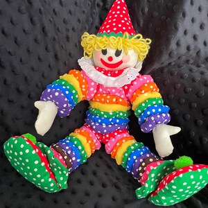 Puede incluir: Una mu&ntilde;eca payaso colorida con un sombrero de lunares rojos y blancos y zapatos a juego. El cuerpo del payaso est&aacute; hecho de tela de colores del arco&iacute;ris con lunares blancos. La mu&ntilde;eca tiene un cuello con volantes blanco y pelo de hilo amarillo.