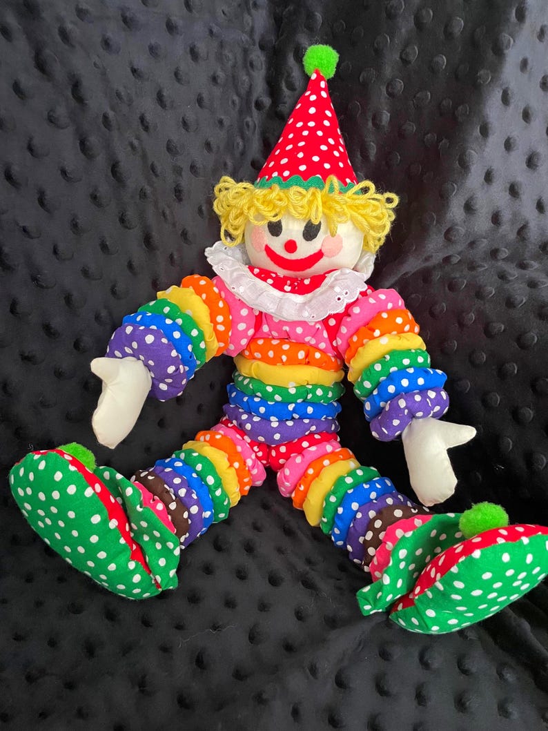 Puede incluir: Una mu&ntilde;eca payaso colorida con un sombrero de lunares rojos y blancos y zapatos a juego. El cuerpo del payaso est&aacute; hecho de tela de colores del arco&iacute;ris con lunares blancos. La mu&ntilde;eca tiene un cuello con volantes blanco y pelo de hilo amarillo.