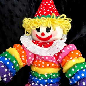 Puede incluir: Una mu&ntilde;eca payaso colorida con un sombrero de lunares rojos y blancos y un atuendo a juego. El payaso tiene un cuello blanco con volantes, cabello de hilo amarillo y una cara sonriente. Los brazos y el cuerpo del payaso son de tela arco&iacute;ris.