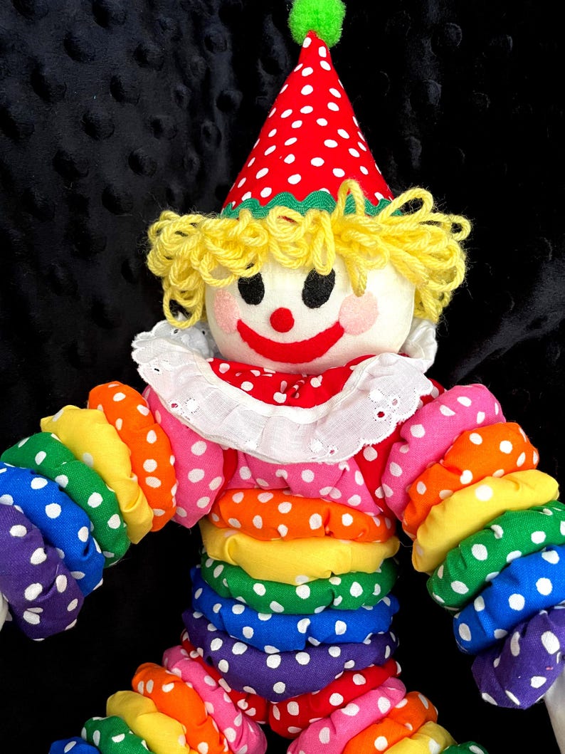 Puede incluir: Una mu&ntilde;eca payaso colorida con un sombrero de lunares rojos y blancos y un atuendo a juego. El payaso tiene un cuello blanco con volantes, cabello de hilo amarillo y una cara sonriente. Los brazos y el cuerpo del payaso son de tela arco&iacute;ris.