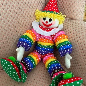 Puede incluir: Mu&ntilde;eca de payaso colorida con sombrero de lunares rojos y blancos y zapatos a juego. La mu&ntilde;eca tiene una cara blanca con una sonrisa roja y cabello de hilo amarillo. El atuendo del payaso es un arco&iacute;ris de colores con lunares blancos.