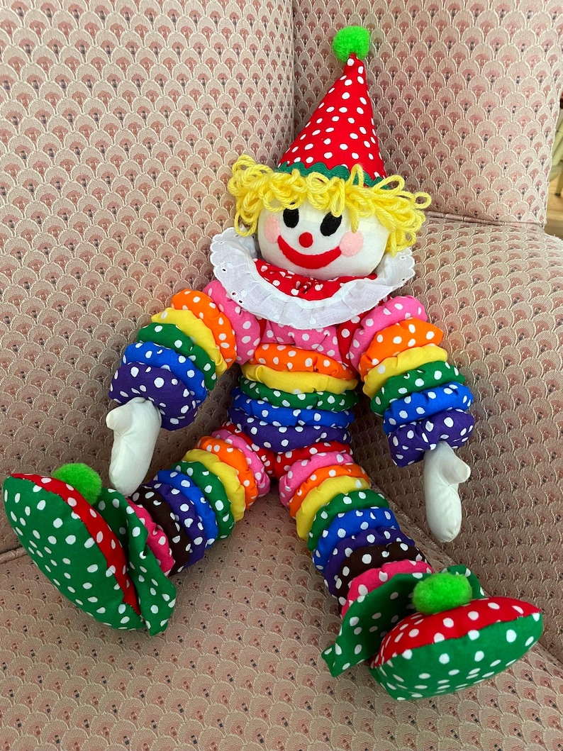 Puede incluir: Mu&ntilde;eca de payaso colorida con sombrero de lunares rojos y blancos y zapatos a juego. La mu&ntilde;eca tiene una cara blanca con una sonrisa roja y cabello de hilo amarillo. El atuendo del payaso es un arco&iacute;ris de colores con lunares blancos.