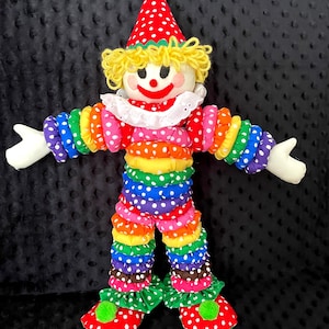 Puede incluir: Mu&ntilde;eca de payaso colorida con sombrero de lunares rojos y blancos y zapatos a juego. El payaso tiene una cara blanca, pelo de hilo amarillo y una sonrisa roja. El cuerpo est&aacute; hecho de tela apilada y con volantes en colores arco&iacute;ris.