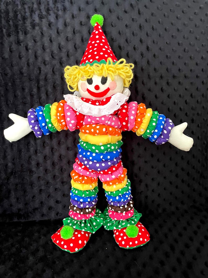 Puede incluir: Mu&ntilde;eca de payaso colorida con sombrero de lunares rojos y blancos y zapatos a juego. El payaso tiene una cara blanca, pelo de hilo amarillo y una sonrisa roja. El cuerpo est&aacute; hecho de tela apilada y con volantes en colores arco&iacute;ris.