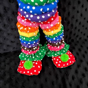Puede incluir: Primer plano de las piernas y los pies de una mu&ntilde;eca payaso. Las piernas est&aacute;n hechas de tela apilada y con volantes en varios colores, incluyendo amarillo, verde, azul, morado, rosa, naranja y marr&oacute;n, con lunares blancos. Los pies son rojos con lunares blancos y pompones verdes.