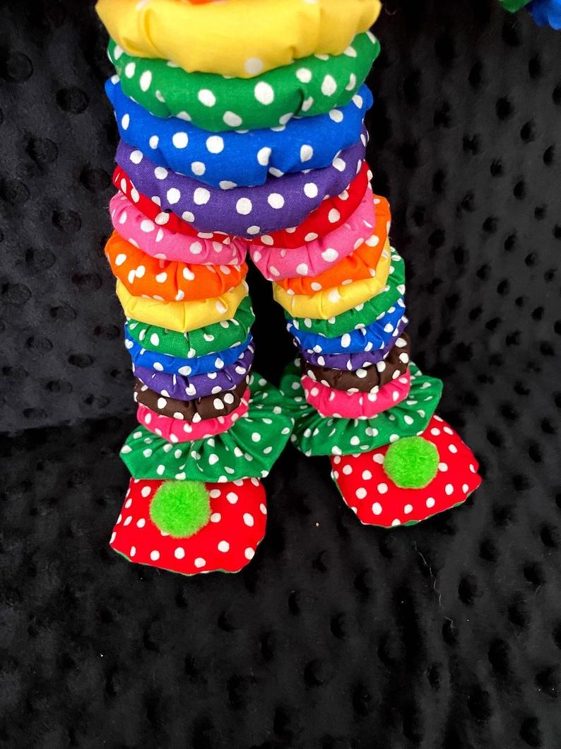 Puede incluir: Primer plano de las piernas y los pies de una mu&ntilde;eca payaso. Las piernas est&aacute;n hechas de tela apilada y con volantes en varios colores, incluyendo amarillo, verde, azul, morado, rosa, naranja y marr&oacute;n, con lunares blancos. Los pies son rojos con lunares blancos y pompones verdes.