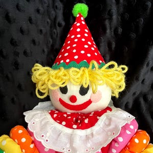 Puede incluir: Mu&ntilde;eca de payaso hecha a mano con sombrero y cuello de lunares rojos y blancos. La mu&ntilde;eca tiene pelo de hilo amarillo, una cara pintada con mejillas rosadas y brazos de tela colorida en tonos rosa, naranja, amarillo y verde.