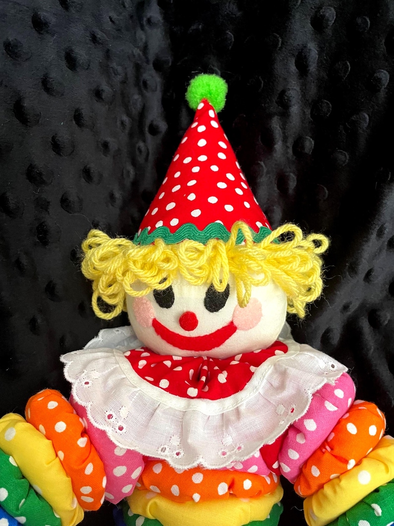 Puede incluir: Mu&ntilde;eca de payaso hecha a mano con sombrero y cuello de lunares rojos y blancos. La mu&ntilde;eca tiene pelo de hilo amarillo, una cara pintada con mejillas rosadas y brazos de tela colorida en tonos rosa, naranja, amarillo y verde.