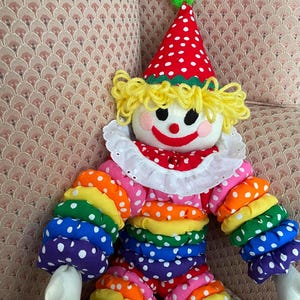 Puede incluir: Mu&ntilde;eca de payaso colorida con un sombrero de lunares rojos y blancos y un pomp&oacute;n verde. La mu&ntilde;eca tiene pelo de hilo amarillo, un cuello blanco con volantes y brazos y piernas de colores del arco&iacute;ris con lunares blancos. La cara del payaso sonr&iacute;e con mejillas rosadas.