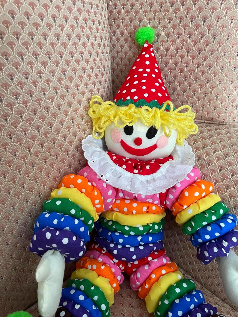 Puede incluir: Mu&ntilde;eca de payaso colorida con un sombrero de lunares rojos y blancos y un pomp&oacute;n verde. La mu&ntilde;eca tiene pelo de hilo amarillo, un cuello blanco con volantes y brazos y piernas de colores del arco&iacute;ris con lunares blancos. La cara del payaso sonr&iacute;e con mejillas rosadas.