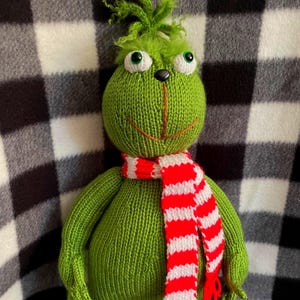 May include: A green knitted Grinch doll with a red and white striped scarf. The doll has large, round eyes, a stitched orange smile, and green hair. The background is a black and white checkered pattern.