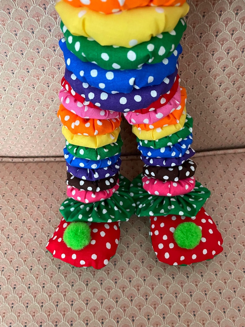 Puede incluir: Piernas de mu&ntilde;eco payaso coloridas. Las piernas est&aacute;n hechas de tela en capas en colores del arco&iacute;ris, cada capa con lunares blancos. Los pies son rojos con lunares blancos y pompones verdes.