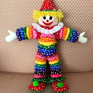 Puede incluir: Mu&ntilde;eca de payaso colorida con sombrero de lunares rojos y blancos y zapatos a juego. El payaso tiene un cuello con volantes blancos, cabello de hilo amarillo y una cara sonriente. Los brazos y las piernas est&aacute;n hechos de tela apilada y con volantes en colores del arco&iacute;ris.