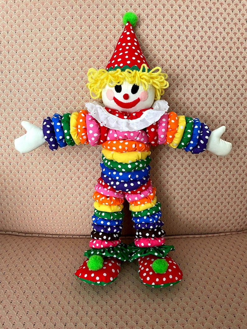 Puede incluir: Mu&ntilde;eca de payaso colorida con sombrero de lunares rojos y blancos y zapatos a juego. El payaso tiene un cuello con volantes blancos, cabello de hilo amarillo y una cara sonriente. Los brazos y las piernas est&aacute;n hechos de tela apilada y con volantes en colores del arco&iacute;ris.