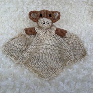 May include: A handmade knitted baby security blanket featuring a monkey face and arms. The monkey's face, ears, and arms are brown, with a cream-colored face and hands. The blanket is cream-colored and has a square shape.